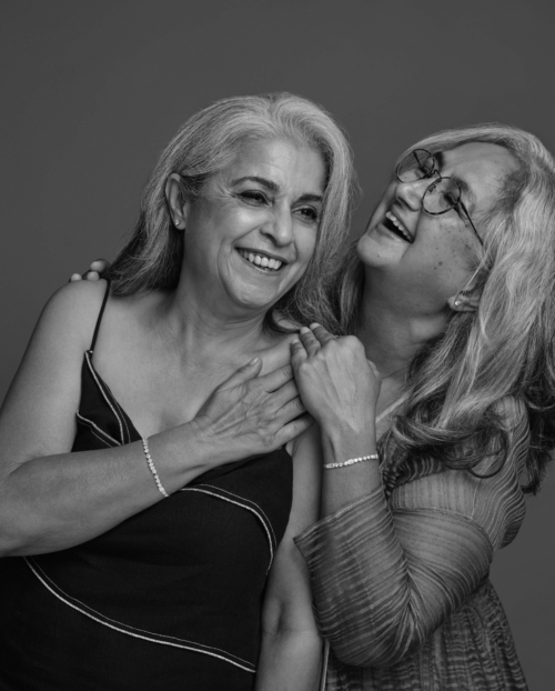 Two older women smiling and laughing together in a joyful moment. One has her hand on her chest, while the other embraces her warmly.