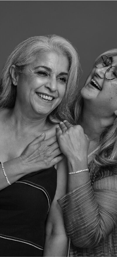 Two older women smiling and laughing together in a joyful moment. One has her hand on her chest, while the other embraces her warmly.