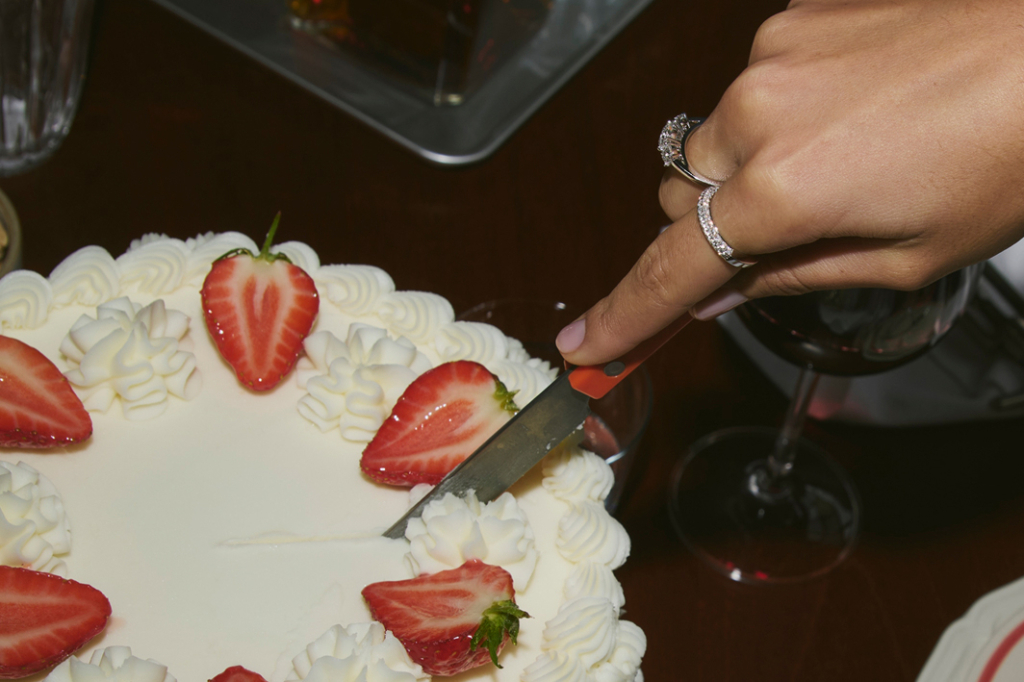 A hand wearing diamond rings cutting a birthday cake