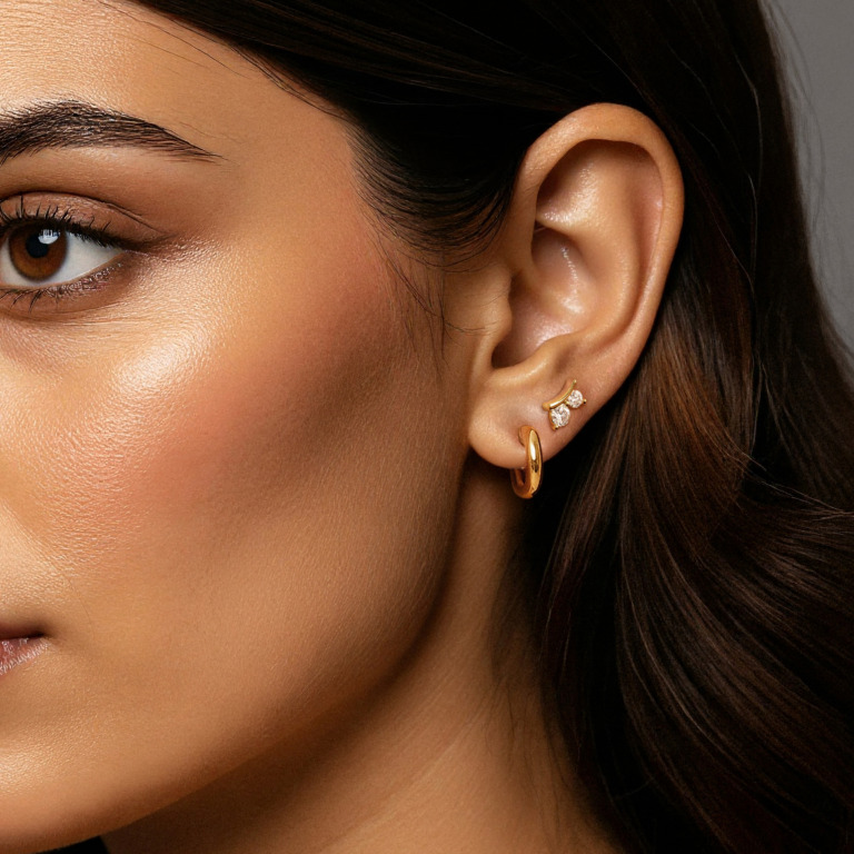 Elegant close‑up portrait featuring a woman with elegant hair in rich brown tones. She has indian features. She is wearing a single statement small gold hoop earring with. Soft, warm studio lighting and a neutral, minimal background. High‑end jewelry editorial style, clean composition, high‑resolution fashion photography.