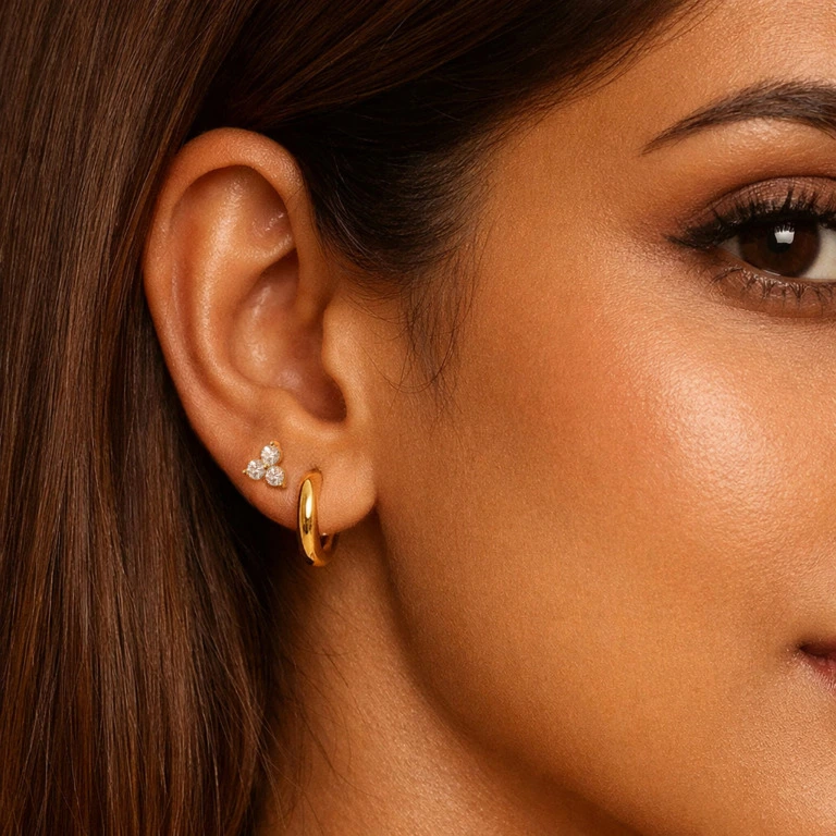 Close‑up aesthetic shot of a woman's ear adorned with a small gold hoop. The woman has indian features. We can see a bit of her eye. Smooth, straight brown hair framing the ear. Soft studio lighting, warm tones, minimalistic and luxurious style, high‑resolution beauty photography, sharp focus on jewelry and texture