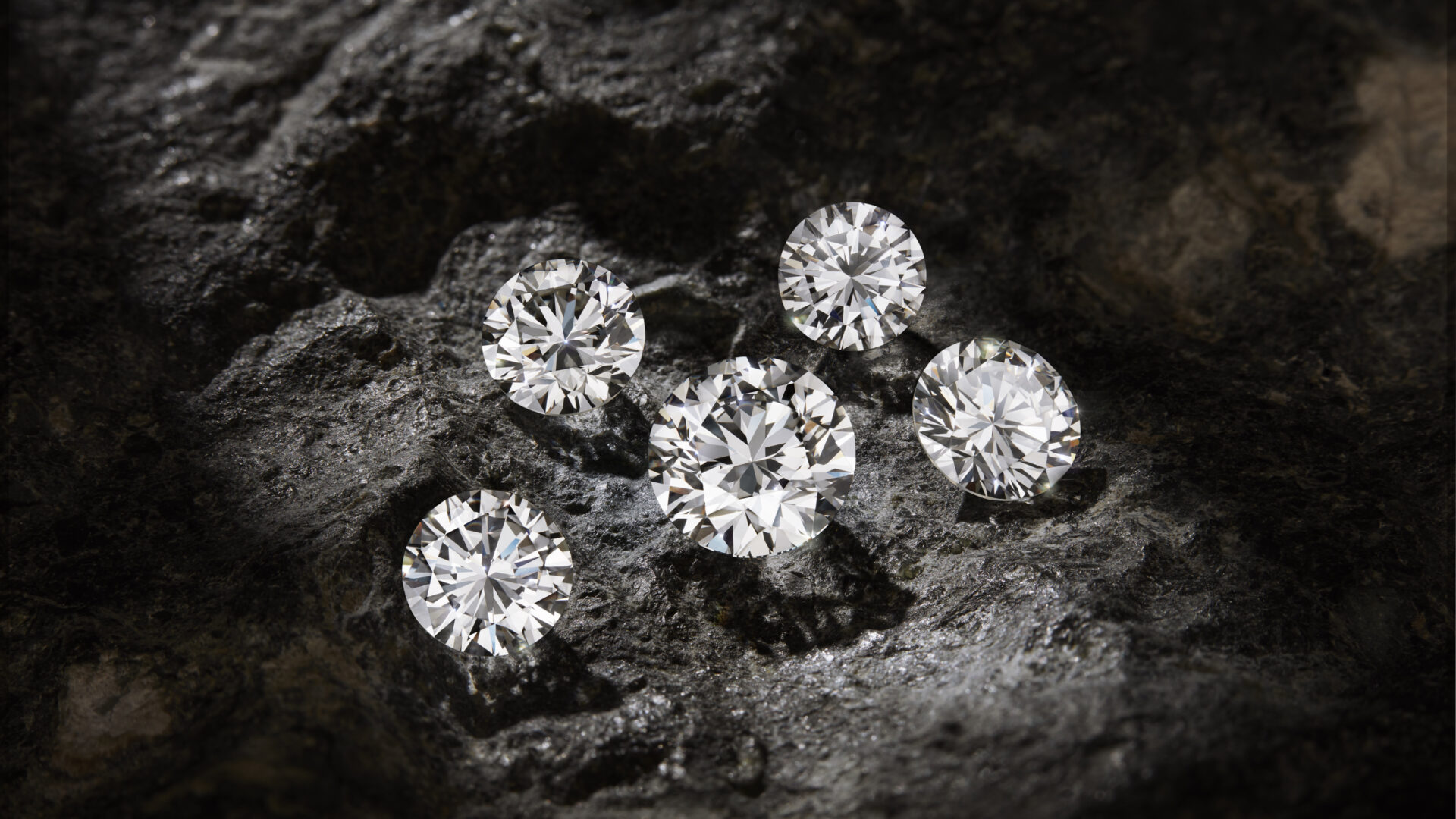 Five brilliant round-cut natural diamonds resting on a dark textured rock surface.