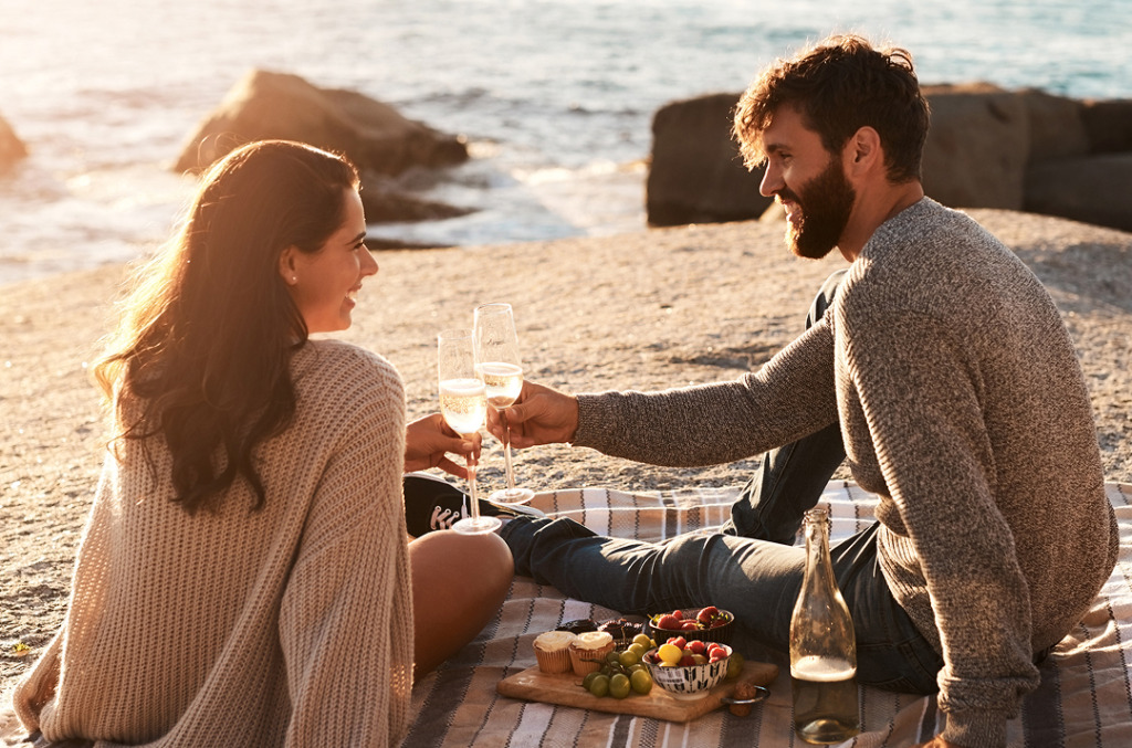 A couple enjoying a seaside picnic at sunset, toasting with champagne as golden light reflects the warm tones reminiscent of Champagne Desert diamonds.