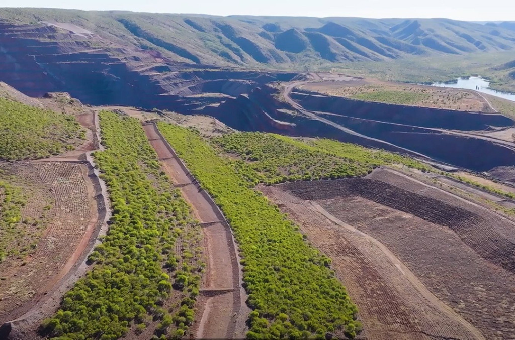 Aerial view of rehabilitated mine land with terraced earth and new vegetation growing across the landscape, set against distant mountains and a winding river.