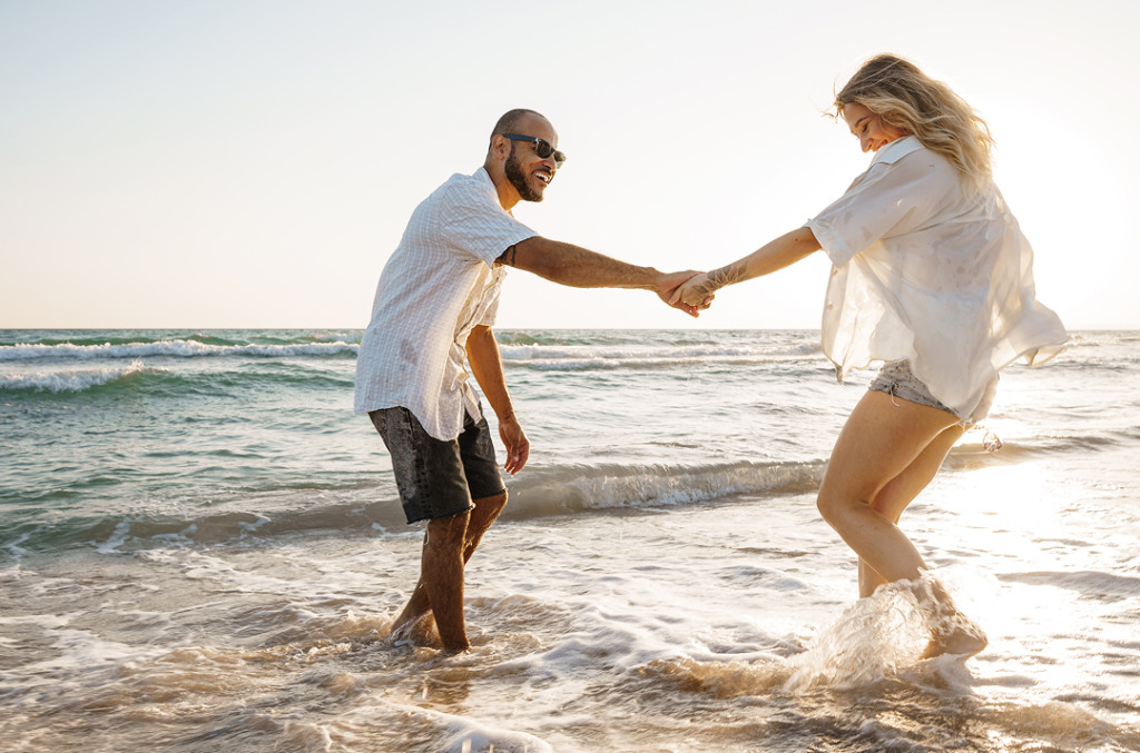 A couple laughing and holding hands while splashing in the waves at the beach during sunset, capturing a playful and romantic moment.