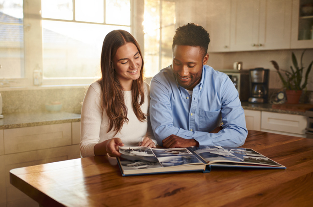 A couple sitting at a kitchen table, smiling as they look through a scrapbook filled with photos and memories.