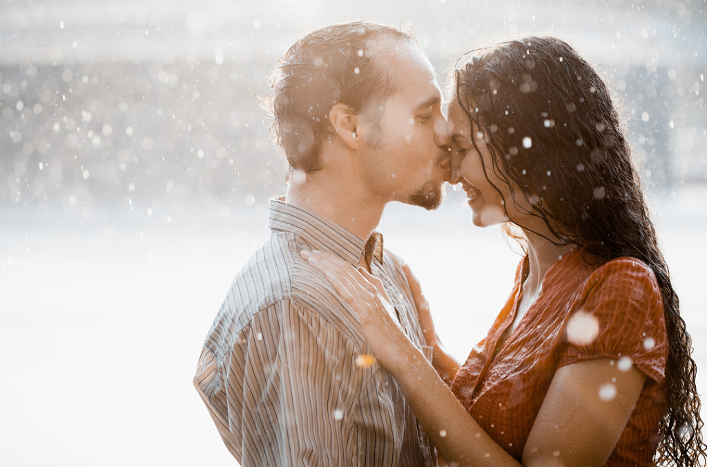 A couple kisses in the rain, smiling with wet hair and clothes, capturing a joyful, unplanned moment that reflects a spontaneous, heartfelt connection.