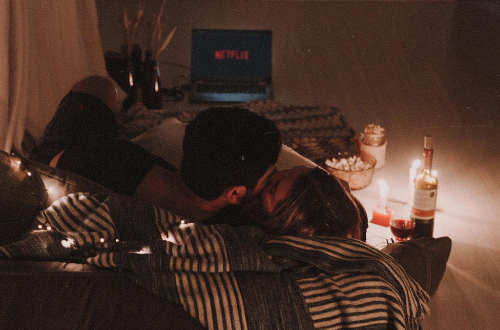 A couple cuddles on the floor surrounded by candles, blankets, wine, and popcorn as they watch a movie at home, creating a warm and intimate setting.