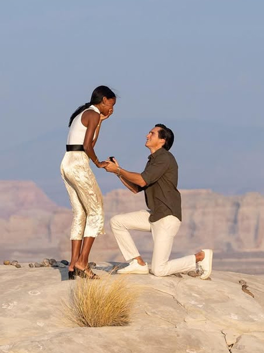 Jasmine Tookes’ fiancé proposing on one knee atop a rocky Utah cliff as she leans forward in surprise and joy.