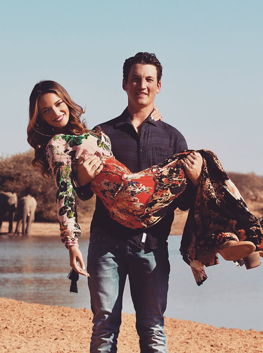 Miles Teller carrying Keleigh Sperry in his arms near a watering hole with elephants in the background during their safari engagement.