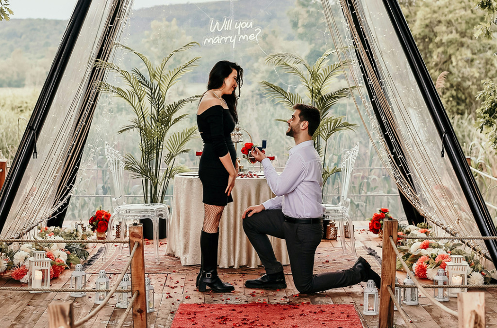 A man kneels to propose in a softly decorated setting with flowers, candles, and a “Will you marry me?” sign as his partner smiles, creating an intimate, sentimental atmosphere.