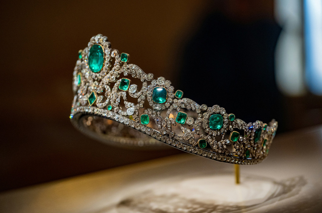 An ornate royal tiara set with large emeralds and intricate arrangements of natural diamonds, displayed on a stand in a museum-like setting.