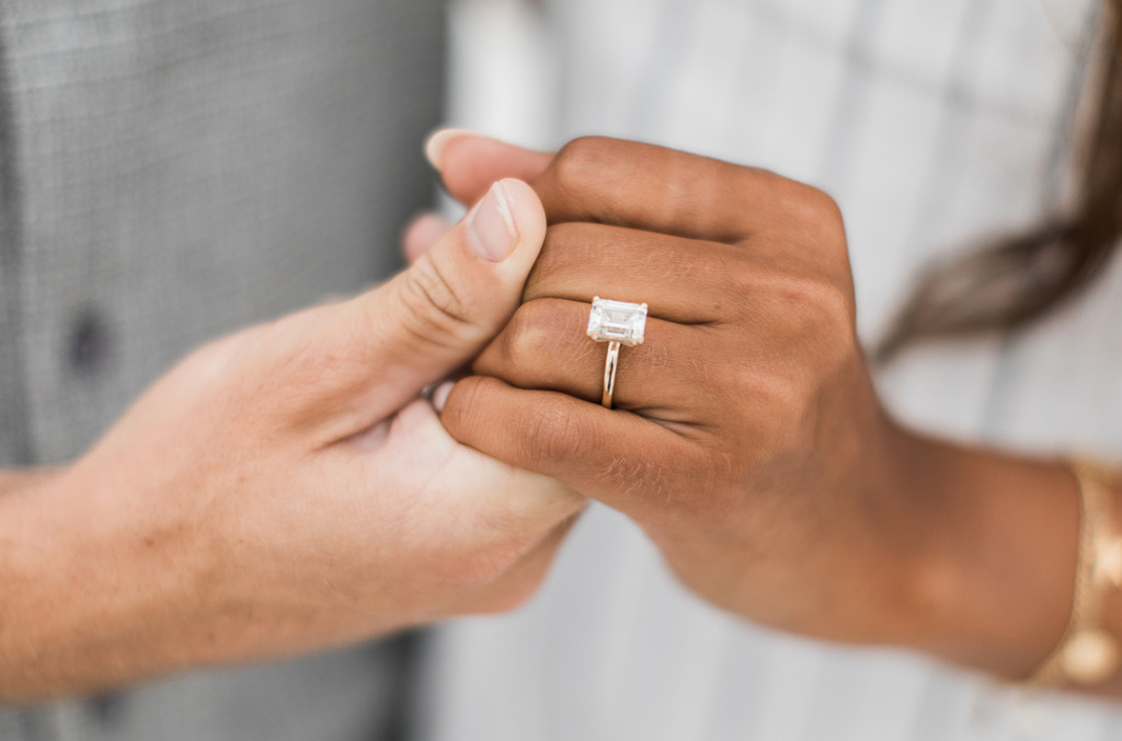 Close-up of two hands intertwined, highlighting an emerald-cut natural diamond ring in a yellow gold setting.