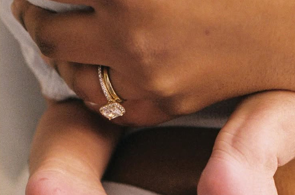 Close-up of a parent’s hand cradling a baby’s foot, wearing a gold cushion-cut diamond ring with a halo setting.