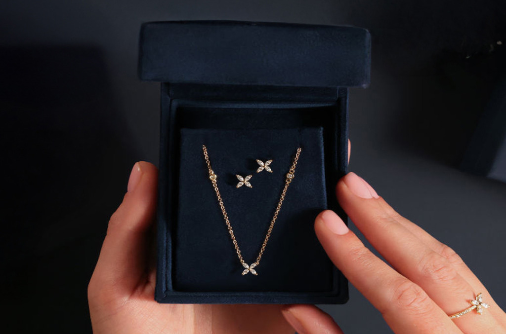 Hands opening a navy velvet box containing a delicate diamond necklace and matching floral-inspired diamond stud earrings.