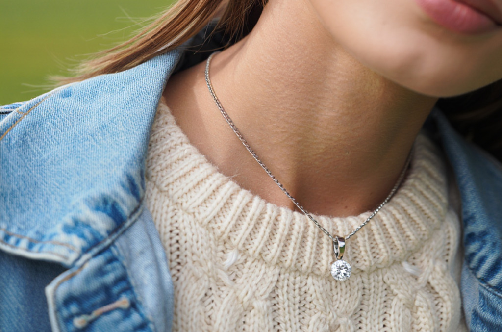 Close-up of a woman wearing a round diamond solitaire pendant on a silver chain, styled with a cream knit sweater and denim jacket.