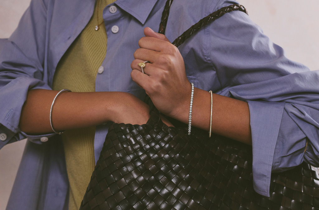 A close-up of a woman holding a woven handbag while wearing natural diamond bracelets and a yellow gold diamond ring.