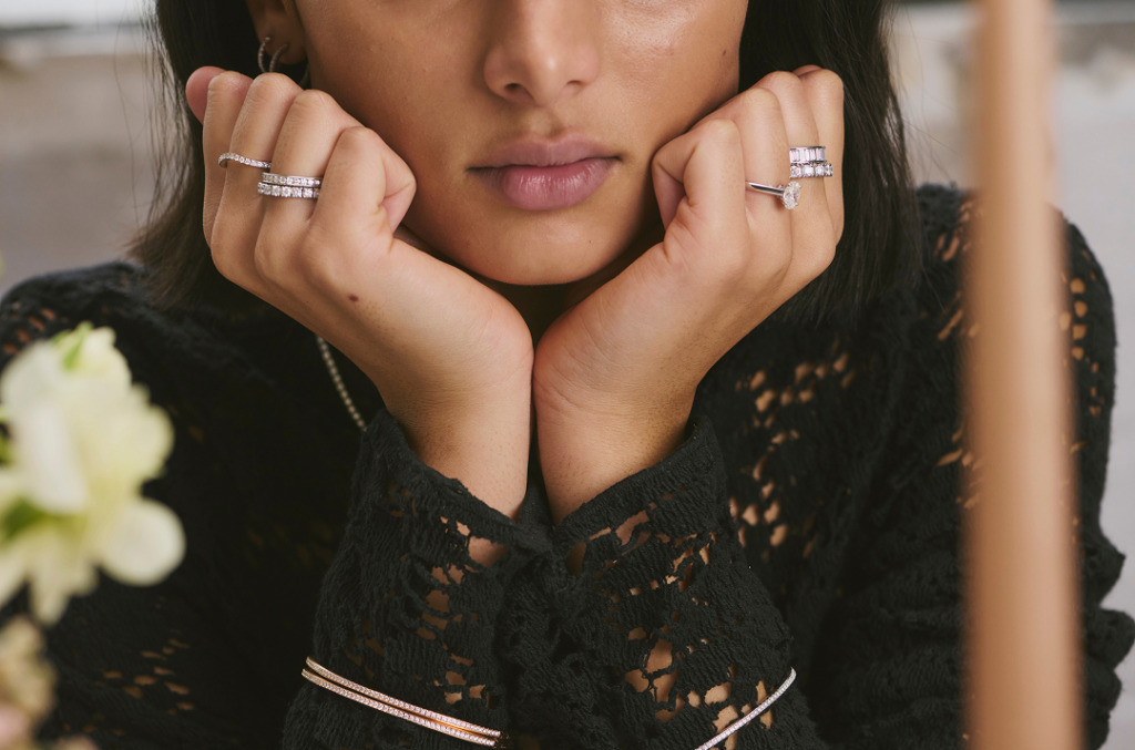 A close-up of a woman resting her chin on her hands, wearing multiple natural diamond rings and bracelets.