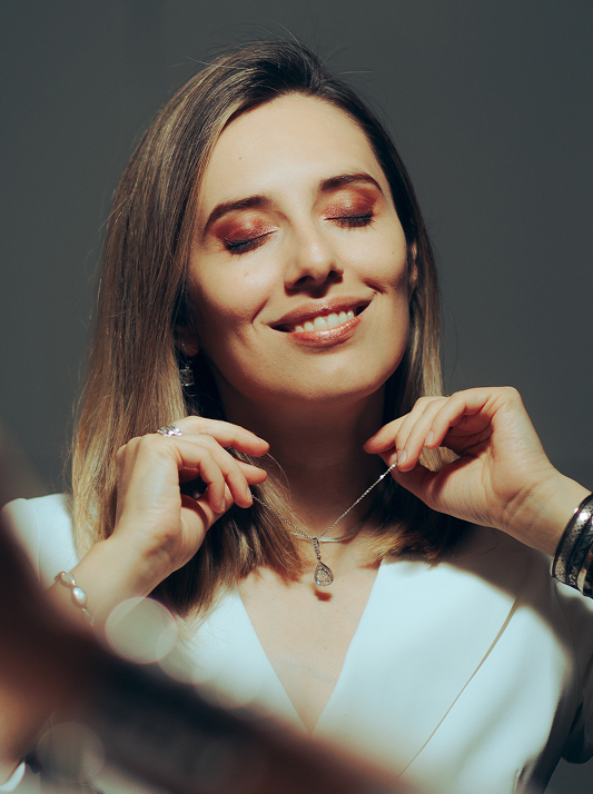 A woman smiling with her eyes closed while clasping a natural diamond pendant around her neck.