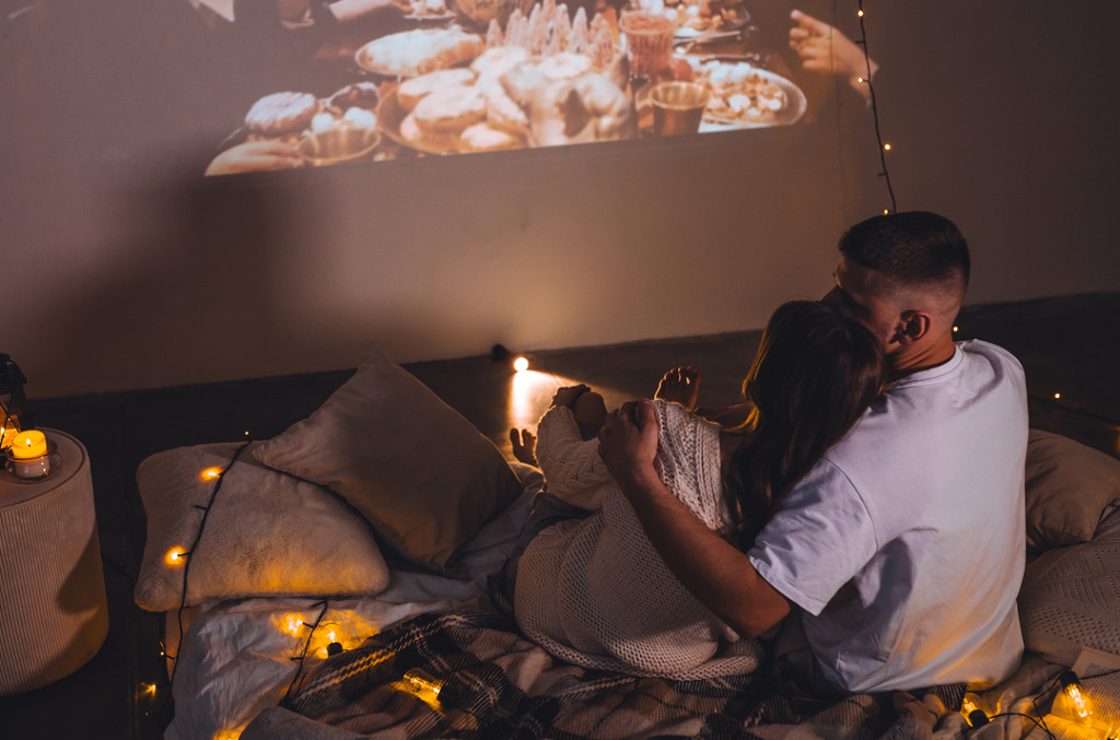 A couple cuddled up on the floor surrounded by blankets and warm string lights, watching a movie projected onto the wall.