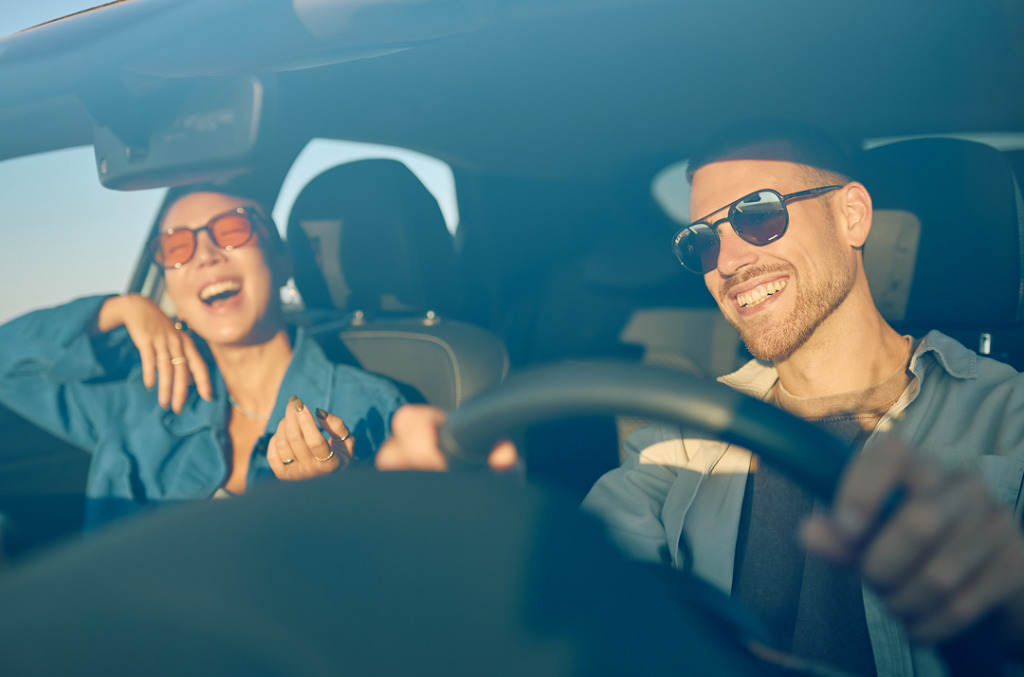 A couple laughing together in a sunlit car, sharing an easy, joyful moment during a drive.