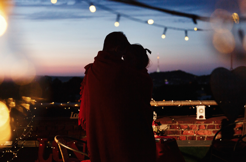 A couple wrapped in a blanket embraces on a rooftop at sunset, surrounded by soft string lights and a warm, romantic glow.