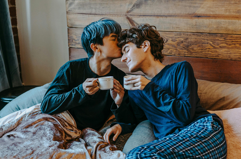 A couple sharing a quiet morning moment in bed, holding coffee mugs as one partner kisses the other’s forehead