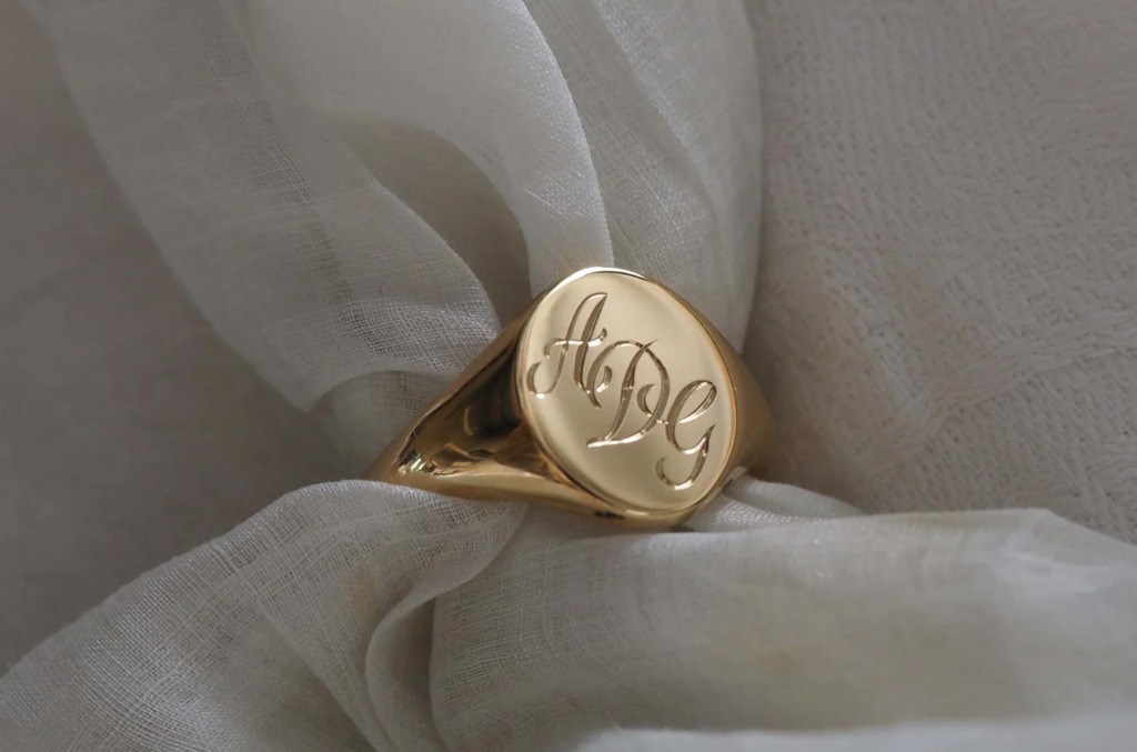 Gold signet ring engraved with intertwined initials, resting on soft fabric, showing how names and letters are engraved into personalized jewelry.
