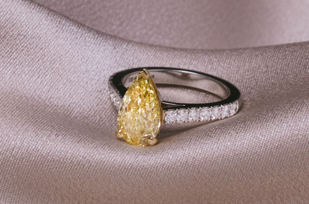 Pear-shaped fancy yellow natural diamond engagement ring with pavé-set white diamonds on a white gold band, photographed on soft fabric.