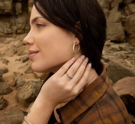 Woman wearing gold hoop earrings and diamond rings, posing outdoors against a rocky background